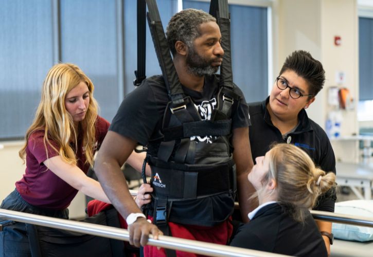 Ray Trowell stands between two railings with assistance from PT students and Jeanette “J.” Lanoire ’17 DPT, a neurologic clinical specialist at Cressman Neurological Rehabilitation.