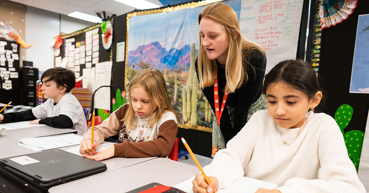 A teacher looks over the work of three students as they write.